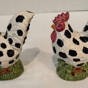 May include: A pair of ceramic rooster figurines. Each rooster is white with black spots, a red comb, and a yellow beak. They stand on a green base with a grass-like texture. Decorative kitchenware.