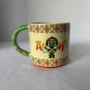 May include: A ceramic mug with a light yellow background and a green handle decorated with flowers. The mug features the word "ALOHA" in colourful letters and a cartoon tiki figure. The rim and base have a decorative pattern.