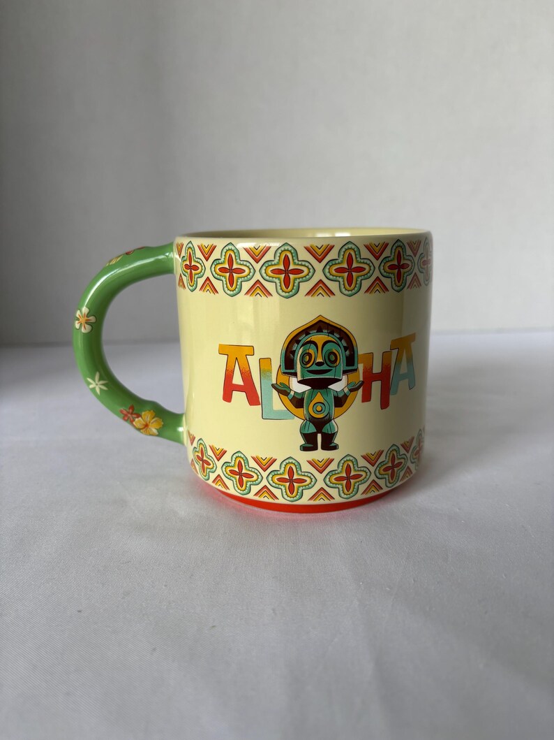 May include: A ceramic mug with a light yellow background and a green handle decorated with flowers. The mug features the word "ALOHA" in colourful letters and a cartoon tiki figure. The rim and base have a decorative pattern.