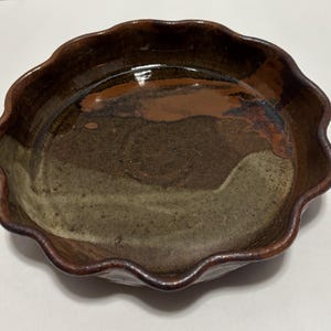 May include: A handmade ceramic pie dish with a wavy rim. The dish features a blend of brown, tan, and olive green hues, creating a rustic and earthy aesthetic. The interior has a glossy finish, while the rim has a darker, matte appearance.