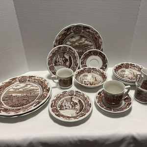 May include: A collection of brown and white ceramic dinnerware, including plates, bowls, saucers, and cups. Each piece features a detailed landscape scene within a decorative border. The set includes various sizes and shapes, suitable for a complete table setting.
