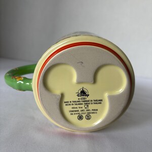 May include: A yellow ceramic mug with a green handle and red trim. The base of the mug features a Mickey Mouse head shape and the Disney logo. The mug is made in Thailand and holds 450 ml.