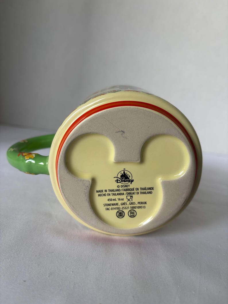 May include: A yellow ceramic mug with a green handle and red trim. The base of the mug features a Mickey Mouse head shape and the Disney logo. The mug is made in Thailand and holds 450 ml.