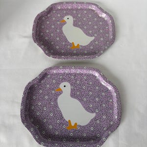 May include: Two decorative trays with a scalloped edge. Each tray features a white duck with an orange beak and feet on a lavender background with a floral pattern. The trays are oval-shaped.
