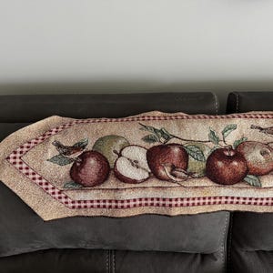 May include: A rectangular tapestry table runner featuring a repeating pattern of apples, leaves, and birds. The runner has a beige background with a red and white checkered border and a brown tassel on one end. The apples are red and green.