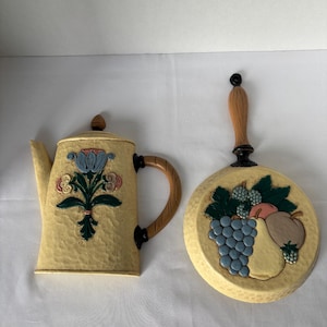 May include: Cream-colored decorative kitchen wall art set. Includes a teapot with a floral design and a round pan with a fruit arrangement. Both pieces have textured surfaces and wooden handles.