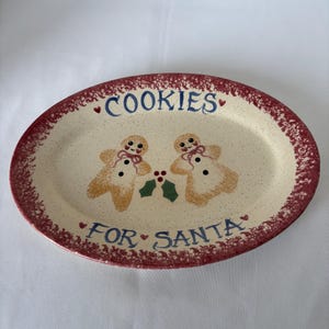May include: An oval ceramic platter with a cream-colored center and a speckled red rim. The text "COOKIES FOR SANTA" is written in blue. Two gingerbread figures with pink bows and black buttons are centered with a green holly sprig and red berries.