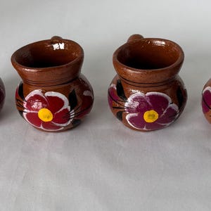 May include: Four small, brown ceramic mugs with handles, each adorned with a hand-painted floral design. The flowers feature a vibrant combination of red, purple, yellow, and white, adding a pop of color to the rustic pottery.