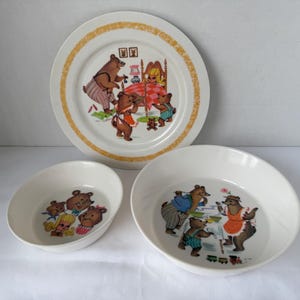 May include: A set of three vintage children's dishes featuring a bear family illustration. The set includes a large plate with a gold rim and two bowls. The illustrations depict bears in various scenes, with bright colors and a retro style.