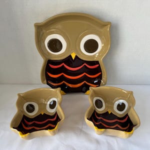May include: A set of three ceramic owl-shaped dishes consisting of one large platter and two smaller bowls. Each piece features a tan face with large white and brown eyes, a yellow beak, and a dark brown body with orange and red scalloped patterns.