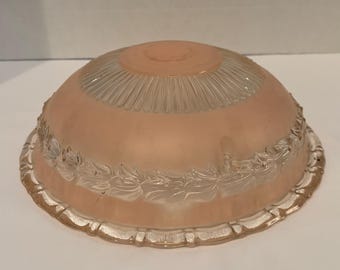 May include: A vintage, frosted glass light fixture shade. The shade is a soft peach color with a clear, ribbed top and a decorative floral pattern around the bottom edge. The shade has a scalloped edge.