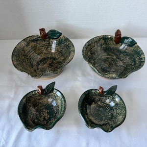 May include: Four ceramic apple-shaped bowls. The bowls are dark green with a speckled, textured pattern and a brown and green stem and leaf detail. The bowls vary in size and are arranged on a white surface.