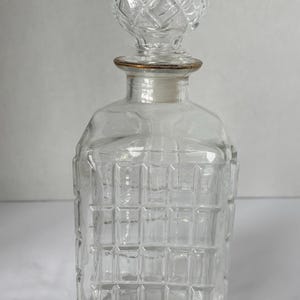 May include: A clear glass decanter with a square body and a decorative, diamond-patterned stopper. The decanter has a gold-colored rim around the neck. The body of the decanter has a grid-like pattern.
