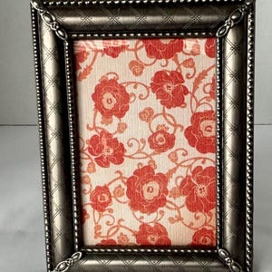 May include: A rectangular, antique silver picture frame with a floral print background. The frame has a decorative, textured border and ornate corner details. The floral print features red flowers on a cream background.