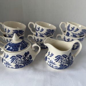 May include: A set of cream-colored ceramic tea service pieces featuring a blue willow pattern. The collection includes five tea cups, a lidded sugar bowl with two side handles, and a creamer pitcher with a pouring spout and a single handle.