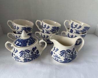 May include: A set of cream-colored ceramic tea service pieces featuring a blue willow pattern. The collection includes five tea cups, a lidded sugar bowl with two side handles, and a creamer pitcher with a pouring spout and a single handle.