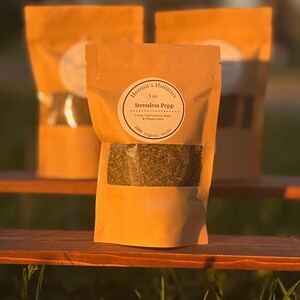 May include: Three brown paper bags of herbal tea are displayed, with the front bag in focus. The bags have a clear window showing the tea leaves. The label reads "Hermit's Holistics" and "Stressless Pepp" with "100% organic herbs."