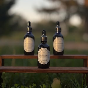 Può includere: Tre bottiglie di vetro blu scuro di olio di origano con contagocce neri sono disposte su un piccolo ripiano di legno. La bottiglia centrale ha un'etichetta che recita "Hermit's Holistic Oil of Oregano 2 oz". Lo sfondo è sfocato con fogliame verde.