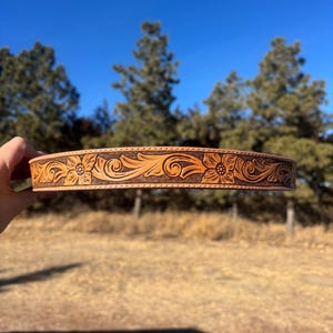 Pode incluir: Um cinto de couro castanho claro com um design floral e de rolagem. O cinto apresenta flores em relevo e padrões giratórios. O fundo mostra um céu azul e árvores.