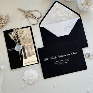 May include: Elegant black wedding invitation suite with a floral design on the envelope liner. Includes a black invitation card with dried flowers, a silver wax seal, and a matching envelope. The suite also includes a black envelope with the recipient's address and a pair of wedding rings.