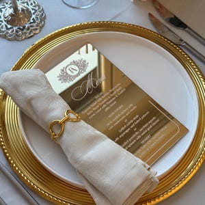 Könnte beinhalten: Eine formelle Tischdekoration mit einem goldgeränderten Teller, einem weißen Teller und einem goldenen Serviettenring. Eine goldene Menükarte mit dem Wort "Menü" ist sichtbar. Eine gefaltete weiße Serviette liegt auf dem Teller. Besteck und ein Weinglas sind ebenfalls vorhanden.