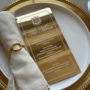 以下が含まれることがあります： 「The Menu」の文字と料理のリストが書かれた金色のメニューカード。金色の縁取りのある白いお皿の上に置かれています。折りたたまれた白いナプキンは、金のリングで留められています。