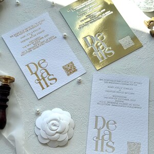 Puede incluir: Tres tarjetas de detalles de boda con l&aacute;mina dorada, plateada y estampado blanco. Las tarjetas presentan la palabra "Details" en una fuente estilizada. Las tarjetas incluyen informaci&oacute;n de la ceremonia de boda y un c&oacute;digo QR. Tambi&eacute;n se ve una flor de papel blanco y perlas.