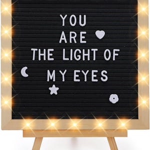 May include: A black felt letter board with the message "YOU ARE THE LIGHT OF MY EYES" in white letters. The board is framed in light wood and has a string of warm white lights around the edge. Decorative shapes include a heart, moon, star, and flower.