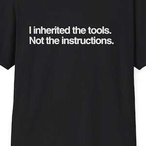 May include: Black t-shirt with white text that reads, "I inherited the tools. Not the instructions." The shirt has short sleeves and a crew neck. The website DeadParents.com is printed at the top.