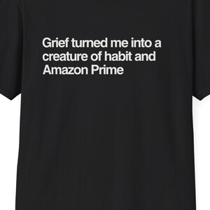 May include: Black t-shirt with the text "Grief turned me into a creature of habit and Amazon Prime" in white. The shirt has short sleeves and a crew neck. The website DeadParents.com is printed at the top.