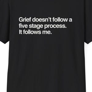 May include: Black t-shirt with the text "Grief doesn't follow a five stage process. It follows me." in white. The shirt has short sleeves and a crew neck.