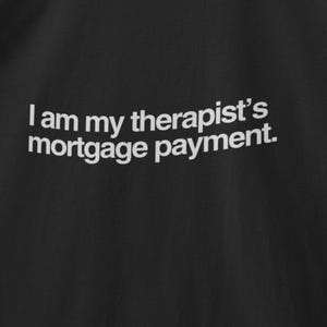 May include: Black t-shirt with the white text: "I am my therapist's mortgage payment." The shirt is hanging on a wooden hanger against a gray background. The t-shirt has a crew neck and short sleeves.