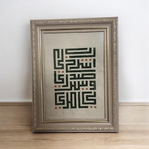 Puede incluir: Obra de arte en punto de cruz enmarcada con caligrafía árabe en hilo verde oscuro y óxido. La obra se exhibe en un marco plateado ornamentado, apoyada contra una pared blanca y un suelo de madera. El diseño es geométrico y abstracto.