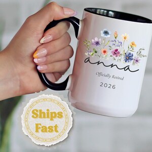 May include: White ceramic mug with a black handle and interior, featuring a floral design and the name "Anna." The mug also includes the text "Officially Retired 2026."
