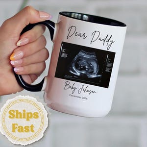 May include: White ceramic mug with a black handle and rim, featuring the text "Dear Daddy" and an ultrasound image. The mug also includes the text "Baby Johnson" and the date "December 2020".
