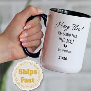 May include: White ceramic mug with a black handle and interior. The mug features the text "Hey Tia! HAZ CAMPO PARA UNO MÁS! NOS VEMOS EN 2026" in black script. The mug is held by a person.