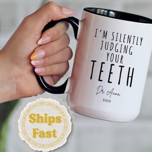 May include: White ceramic mug with a black handle and rim. The mug features the text "I'M SILENTLY JUDGING YOUR TEETH" in black, with "Dr. Anna DDS" below. The mug is held by a hand with manicured nails.