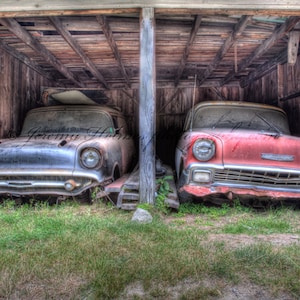 May include: Two vintage cars, one black and one red, are parked inside a wooden garage. The cars are both classic American models with chrome accents and faded paint.