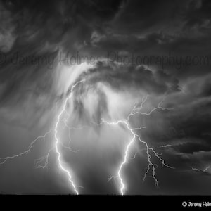 May include: A black and white photograph of a lightning strike in a stormy sky. The lightning bolt is shaped like a heart and is surrounded by dark clouds.