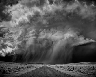 May include: A dramatic black and white landscape photograph of a severe thunderstorm. A dark, ominous cloud formation dominates the sky, with rain falling over a rural road. The image captures the power and intensity of the storm.