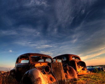 Könnte beinhalten: Zwei alte, rostige und verwitterte Autos stehen auf einem Feld mit hohem Gras. Der Himmel ist tiefblau mit schwebenden Wolken. Das Auto im Vordergrund ist eine klassische Limousine mit einem verchromten Kühlergrill und Scheinwerfern. Das Auto im Hintergrund ist eine größere Limousine mit einer abgerundeten Form.