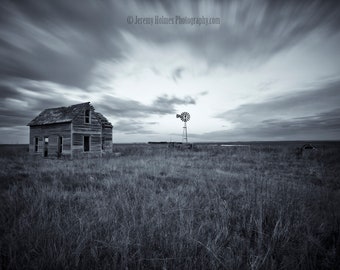 Könnte beinhalten: Ein Schwarzweißfoto eines verwitterten Holzfarmhauses mit einem Windrad in der Ferne. Das Farmhaus ist von hohem Gras umgeben und der Himmel ist bewölkt.