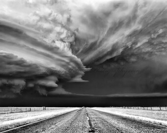 May include: A black and white photograph of a dramatic, swirling storm cloud over a straight, empty highway.