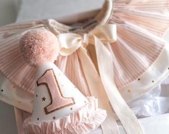 May include: A pink and white party hat with a gold glitter number one and gold stars. The hat has a pink pom-pom on top and a ruffled edge.