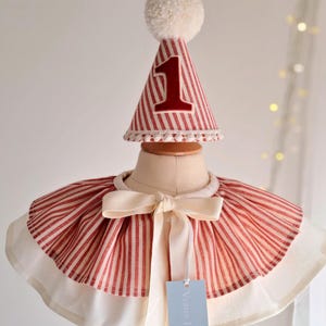 May include: A red and white striped party hat with a large number 1 in red, topped with a white pom-pom. The hat sits above a matching red and white striped cape with a cream-colored bow. A tag is attached to the cape.