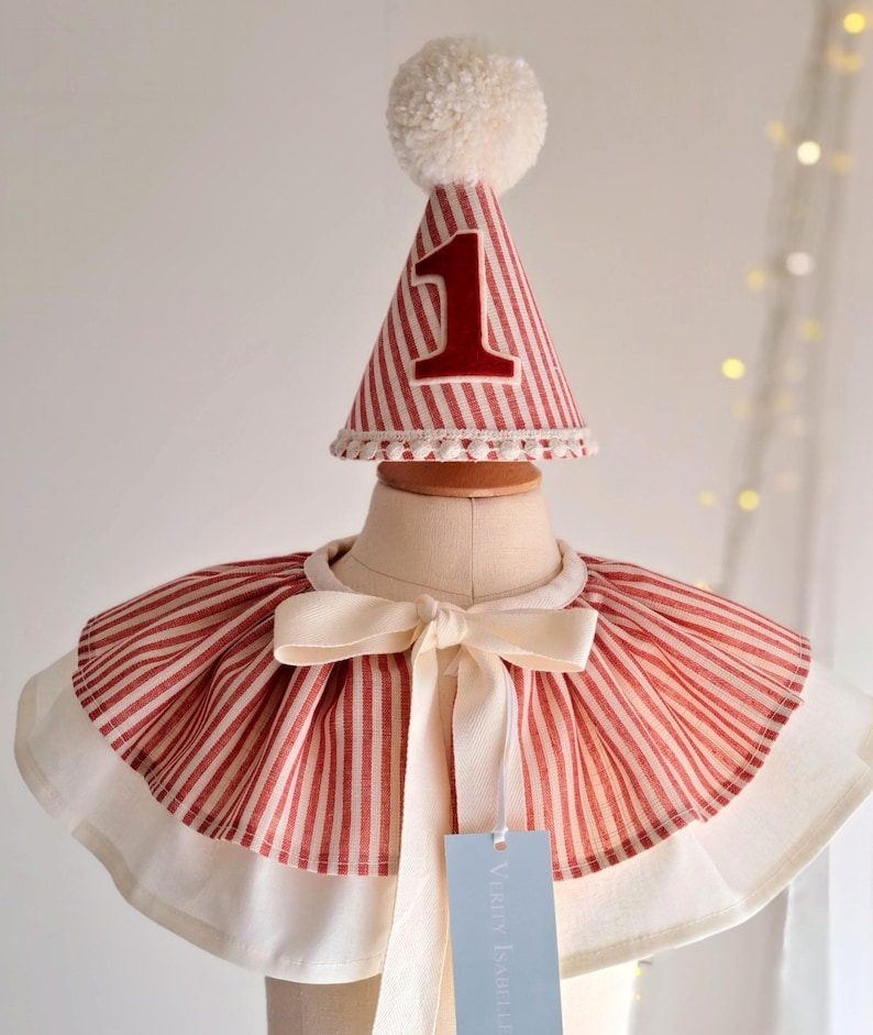 May include: A red and white striped party hat with a large number 1 in red, topped with a white pom-pom. The hat sits above a matching red and white striped cape with a cream-colored bow. A tag is attached to the cape.