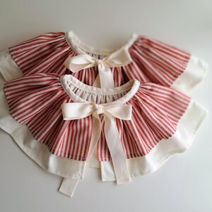 May include: Two red and white striped bibs with cream-colored bows. The bibs have a ruffled edge and a cream-colored border. The bows are tied in the center of the bibs.