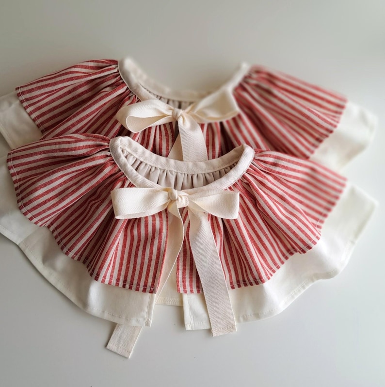 May include: Two red and white striped bibs with cream-colored bows. The bibs have a ruffled edge and a cream-colored border. The bows are tied in the center of the bibs.
