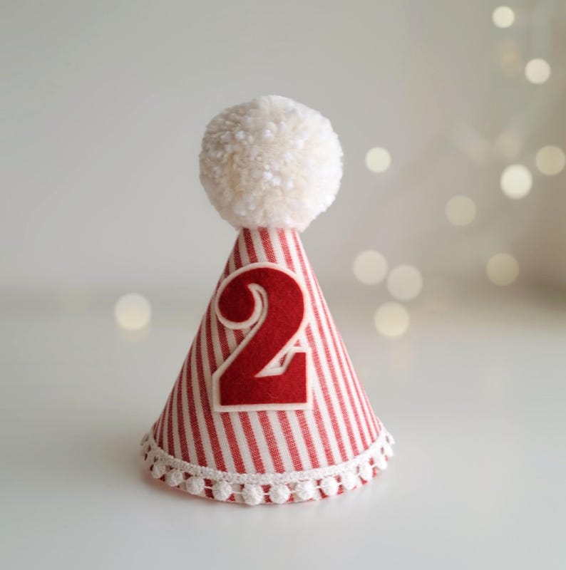 May include: A festive party hat with red and white stripes, a white pom-pom, and a large red number 2. The hat has a white pom-pom trim at the base. Perfect for a second birthday celebration.