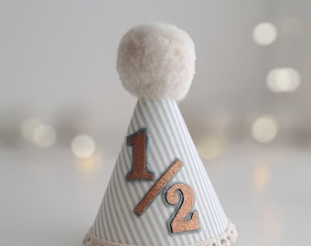 May include: A cone-shaped party hat with gray and white stripes, a beige pom-pom, and a decorative trim. The hat features the numbers "1" and "1/2" in a copper colour. The hat is designed for a half-birthday celebration.